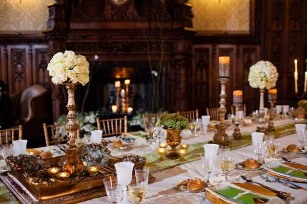 A wedding reception tablescape shot in a classic, formal indoor setting with dark carved wood paneling and an ornate fireplace visible in the background. The long dining table is dressed with a cream linen base and a green runner, set with crystal glassware, gold-toned charger plates, green folded napkins, and individual bread rolls. Tall gold candelabra centerpieces hold large spherical arrangements of cream/ivory roses, flanked by pillar candles on gold candlesticks and small gold votive tea light holders arranged on a decorative gold tray. The overall decor palette is gold, cream, and green with a classic, formal aesthetic. Wide shot capturing the full length of the table from a slightly elevated angle. Potential venue feature image.