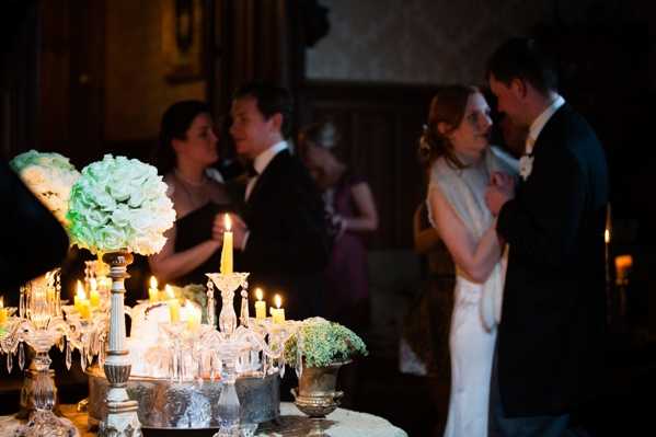 A couple shares a slow dance or intimate moment during an indoor evening reception, with the bride in a white gown and the groom in a dark suit with white bow tie visible in the right foreground. Several guests, including a woman in a strapless dark gown and a man in a tuxedo, stand and converse in the background alongside another guest in a purple dress. The foreground features a richly styled tablescape with multiple lit candles on crystal and silver candelabras, small arrangements of what appear to be white hydrangeas and baby's breath in silver urns, and glassware, all creating warm candlelight throughout the dark interior space. The overall decor palette is classic and formal with silver, crystal, and white floral tones, shot in a medium-depth portrait style with shallow focus that keeps the table decor sharp and the background softly blurred.