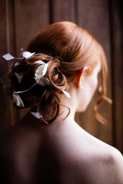 A close-up portrait of a bride shot from behind, showcasing her updo hairstyle. The bride has auburn hair styled into a loose, romantic chignon adorned with small white flower petals or floral hair accessories tucked throughout the bun. She appears to be wearing a strapless or off-shoulder gown, with her bare back visible. The image is a detail/portrait shot with a dark wooden backdrop, creating warm, moody contrast against her hair and skin tone.
