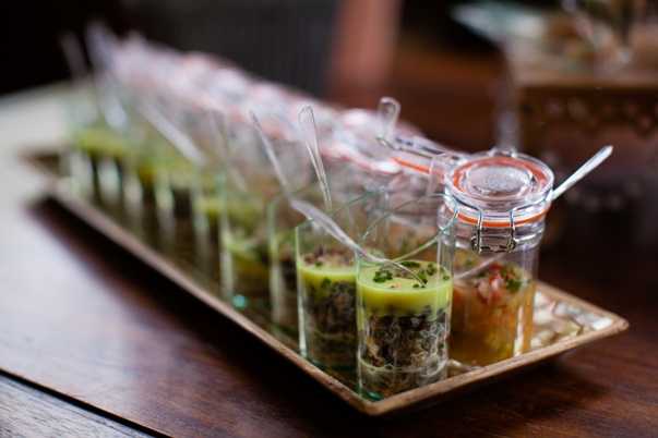 Close-up detail shot of wedding cocktail hour canapés arranged on a rectangular gold/bronze tray placed on a dark wood surface. A row of small clear shot glasses filled with layered appetizers — featuring a yellow-green sauce topped with dark herb mixtures — are each fitted with small clear spoons. To the right, a small hinged-lid glass jar contains a red and pink chopped filling, consistent with a tartare or salsa-style amuse-bouche. The background is softly blurred, with additional food presentations visible out of focus, suggesting an indoor catering or reception setting.