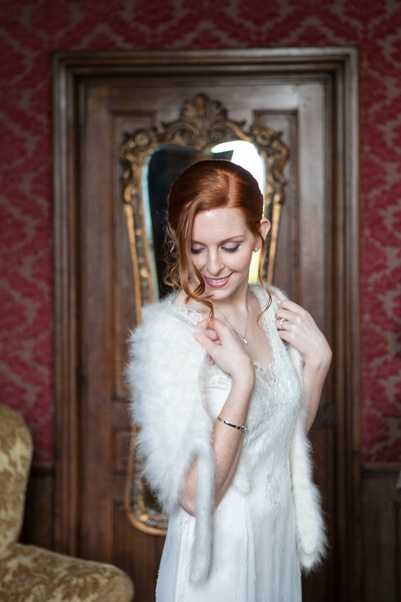 A getting-ready portrait of a bride with red hair, wearing a white lace wedding dress and a white feather or marabou stole draped over her shoulders, paired with a simple white headband. She is smiling and looking downward while adjusting the stole. The setting is an ornate indoor room with deep red damask wallpaper, a carved wooden door, and a gold-framed mirror visible in the background, suggesting a classic chateau or historic venue interior. The shot is a close-up portrait with a shallow depth of field, giving the background a soft blur, and the overall styling leans toward a vintage or classic aesthetic.