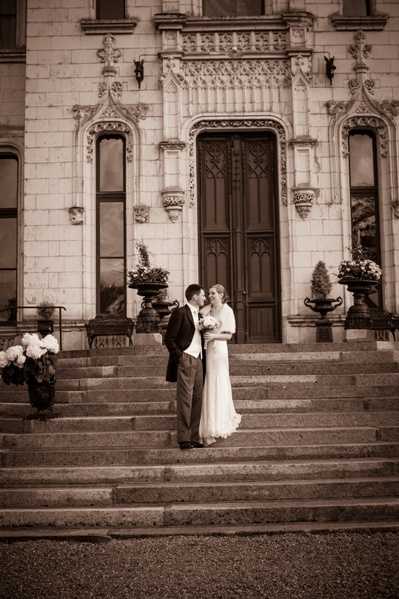 A sepia-toned portrait of a bride and groom sharing a close moment on the stone entrance steps of a French chateau, facing each other with the groom leaning in toward the bride. The bride wears a fitted, long white gown and holds a small bouquet, while the groom wears a dark suit. The chateau facade behind them features ornate Gothic-style carved stonework, arched windows, and a large dark wooden double door flanked by decorative stone urns with floral arrangements and trimmed topiary. The composition is a full-length portrait framing the couple against the grand architectural backdrop. Potential venue feature image.