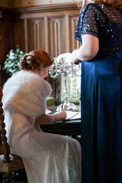 The bride is seated at a dark wooden table signing the wedding register, photographed from behind in a medium portrait shot. She is wearing an ivory wedding dress with a white faux fur stole over her shoulders and has her red hair styled in an updo with small floral hair accessories. A woman in a navy blue sequined-top bridesmaid dress stands beside her, partially cropped at the frame's edge. On the table sits a silver candelabra with crystal droplet details. The interior setting features dark wood paneling, suggesting a historic manor or chateau, and the overall decor palette leans classic and wintry.