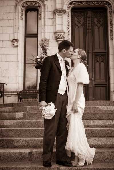 A sepia-toned portrait of a bride and groom kissing on the stone steps of an ornate building with carved architectural details and a large decorative wooden door. The groom wears a dark suit with a light-colored tie, while the bride wears a lace wedding dress with a fur or feather wrap stole over her shoulders. She holds a round bouquet of what appear to be white roses. The composition is a medium full-length shot, capturing both figures centered on the steps with the building facade as backdrop. The sepia processing gives the image warm brown tones with moderate contrast.
