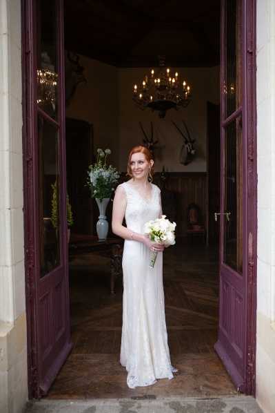 A bride stands centered in the open double doorway of what appears to be a French chateau or manor house, framed by deep plum-purple wooden doors. She wears a fitted, floor-length ivory lace gown with a V-neckline and holds a tight round bouquet of white flowers, likely white calla lilies and roses. Her red hair is styled up and she wears drop earrings; behind her, the interior reveals dark wood paneling, a wrought-iron candle chandelier, animal horn mounts on the wall, and a tall vase of loosely arranged white and green florals. This is a full-length portrait shot taken from slightly outside the doorway, with natural light illuminating the bride against the darker interior.