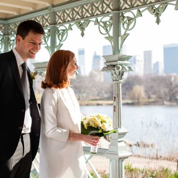 A couple poses together beneath an ornate pale green cast-iron pavilion or gazebo, with a city skyline and body of water visible in the background, suggesting an urban park setting. The bride wears a white long-sleeve coat-style dress and holds a compact round bouquet of white and cream flowers, likely ranunculus or peonies, with green accents and a large leaf wrap. The groom wears a black suit with a white dress shirt, dark tie, and a small white floral boutonniere. The portrait-style shot is taken at mid-distance, capturing both figures from roughly the waist up, with the couple looking at each other and smiling.