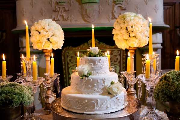 Close-up detail shot of a three-tiered white wedding cake decorated with white fondant scrollwork, white roses, and small green berries, displayed on a gold cake stand. The cake is flanked by crystal candelabras holding lit yellow taper candles, and two large round floral arrangements of cream and white roses on tall gold pedestals. Small arrangements of green moss or foliage are positioned at the base of the display. The backdrop features an ornate carved stone or plaster mantelpiece with decorative molding and a green-and-gold painted interior, suggesting a chateau or historic reception venue interior. The overall decor palette is white, cream, gold, and green with a classic, formal styling.