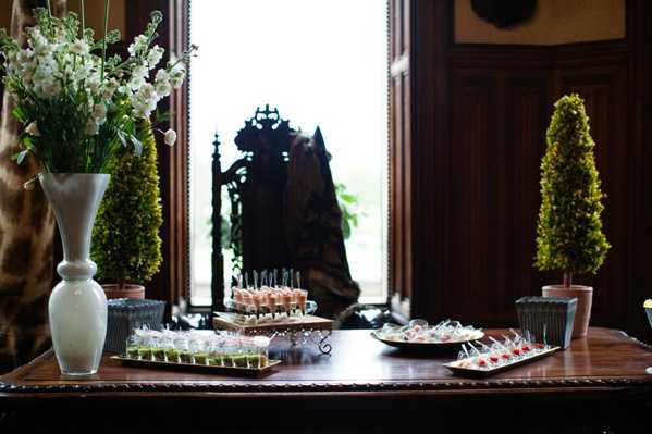 A cocktail hour canapé display table set up indoors in what appears to be a wood-paneled room with dark mahogany wainscoting and a large window in the background. The dark wooden table holds multiple trays and tiered stands of bite-sized appetizers and canapés, including items with pink, green, and red garnishes. The table is styled with a tall white ceramic vase holding white stock flowers on the left, and two small conical boxwood topiary trees in square containers flanking the display. The overall decor palette is classic and formal, using white florals and structured greenery. Close-up wide shot taken at table level.