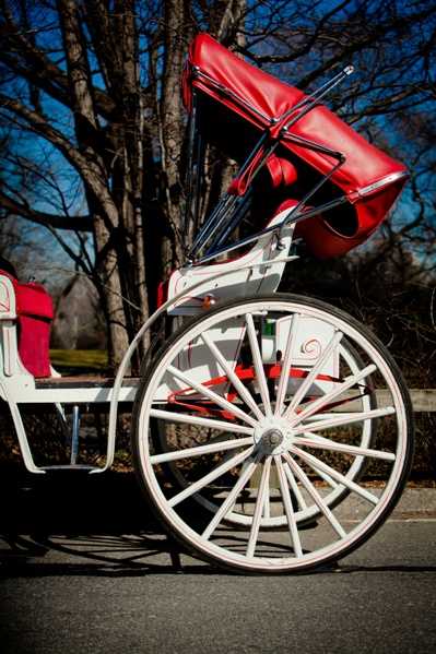 Detail shot of a traditional horse-drawn carriage styled in white and red, with the folded convertible hood and upholstered red leather seating visible. The carriage features large white-painted spoked wheels with decorative scrollwork on the body. No people are visible in the frame. The composition is a close-up side angle focusing on the wheel and carriage body, likely used as a wedding transport detail shot.