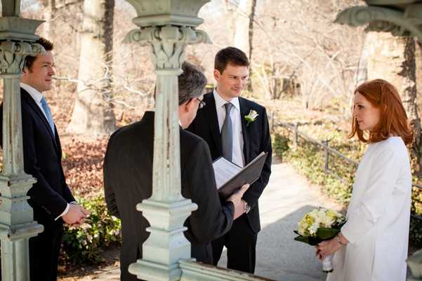 A small outdoor wedding ceremony taking place under a decorative stone pergola or gazebo structure with ornate columns painted in a weathered sage-green color. Four people are present: a groom in a dark navy suit with a white rose boutonniere and grey tie, a bride with red hair wearing a white coat and holding a bouquet of yellow and white flowers, an officiant in a dark suit holding an open binder or ceremony booklet, and a male witness or best man in a dark suit with a light blue tie. The bride and groom are facing each other during what appears to be the vow exchange, with the officiant reading between them. The photograph is a medium-wide shot taken from the side of the pergola, capturing all four participants in a candid, documentary style.