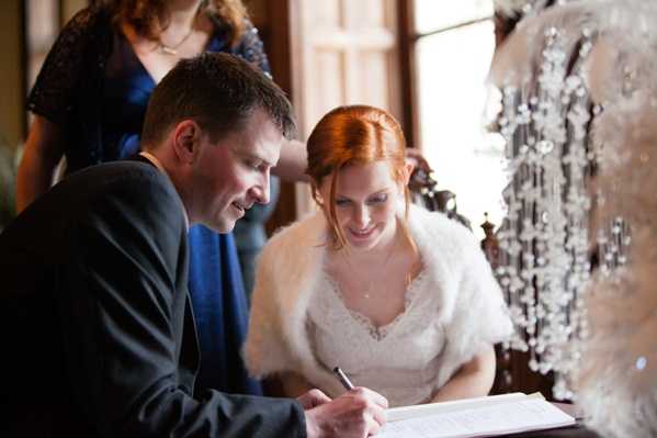 The bride and groom are signing the marriage register during an indoor ceremony, both leaning over an open registry book on a wooden table. The groom wears a dark charcoal suit and holds a pen, while the bride, a redhead with her hair pinned up, wears a lace-bodice dress with a white fur wrap over her shoulders and smiles down at the document. A third person, partially visible in the background, wears a cobalt blue dress and appears to be a witness or officiant. To the right of the frame, a decorative crystal or acrylic beaded chandelier or ornament is partially visible, adding a formal interior detail to the scene. The image is a close-up portrait shot with warm, window-sourced natural light.