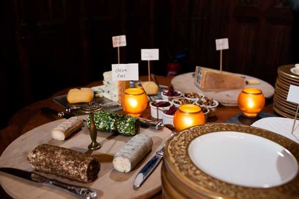 Close-up detail shot of a cheese board display at an indoor wedding reception, likely during a cocktail hour or dinner service. The wooden board features multiple varieties of French cheese including a labeled chèvre frais (fresh goat cheese), a herb-coated log, a cocoa-dusted log, and a wedge of hard cheese, accompanied by small white bowls filled with accompaniments such as fruit preserves and nuts. The table is lit by amber glass votive candles casting warm orange tones, and hand-written label cards identify each cheese variety. Gold-rimmed china plates are stacked in the foreground and to the right, suggesting a formal or classic table setting with warm, candlelit styling.