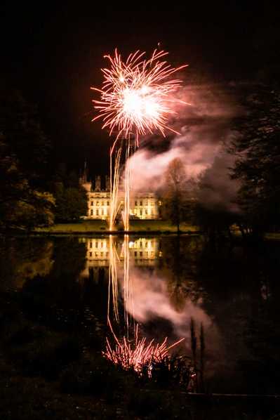 A nighttime fireworks display captured from across a lake, with a large illuminated chateau visible in the background. A single firework bursts in pink and white tones above the building, with the explosion and the lit facade of the chateau reflected in the still water below. The surrounding grounds are dark, with trees framing either side of the composition. The shot is a wide vertical frame emphasizing the symmetry between the firework and its reflection. Potential venue feature image.