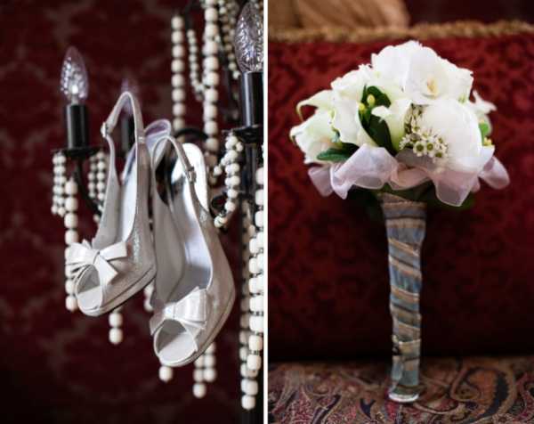 A split close-up detail shot showing two bridal accessories against a richly upholstered deep red background. On the left, a pair of white satin peep-toe slingback heels with bow detail are hung from a black chandelier adorned with cream wooden bead strands. On the right, a bridal bouquet composed of white peonies, white calla lilies, and small white wax flowers with green foliage rests on a red patterned fabric surface; the stems are wrapped in a striped ribbon in muted blue and grey tones, finished with a loose blush pink organza ribbon. The styling has a classic, formal feel with rich jewel-toned surroundings contrasting the white florals and ivory shoes.