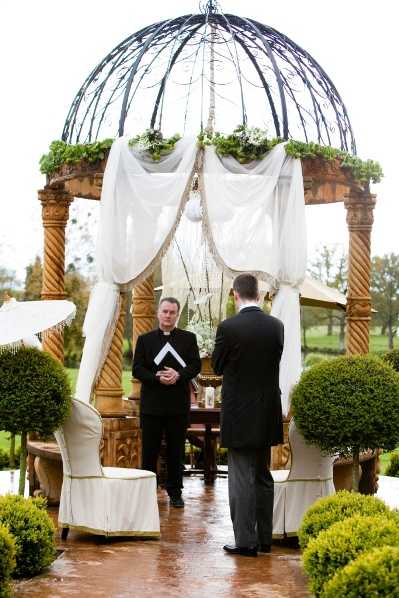 An outdoor wedding ceremony captured in a wide portrait shot, showing a groom in a dark charcoal suit standing before an officiant dressed in a black clerical shirt and holding a ceremony book. The ceremony takes place beneath an ornate dome-shaped gazebo structure with wrought-iron lattice top, twisted gilt columns, and white fabric draping tied at each column. The gazebo is decorated with green foliage and small white flowers along the upper rim, and a lantern is visible inside. Two tall cream-upholstered wingback chairs flank the altar area, and clipped boxwood topiaries are positioned on either side of the aisle. The ground is wet, suggesting the ceremony took place in rainy conditions. The overall decor palette is cream, gold, and green with a classic, formal styling theme.