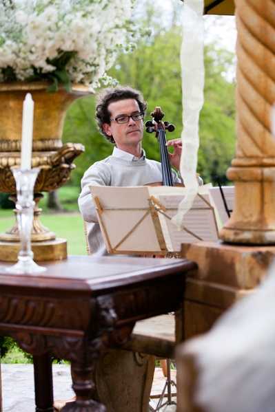 A cellist performs during what appears to be an outdoor wedding ceremony, seated beneath a covered structure with gold-painted columns draped with white ribbon. The musician, a man wearing glasses and a light grey sweater, plays while reading from sheet music on a wooden music stand. In the foreground, an ornate dark wood carved table holds a tall gold candelabra with a white taper candle, and large gold urn arrangements filled with white florals — likely white hydrangeas and small white blooms — are visible to the left. The shot is a medium portrait framing the musician between the decorative columns, with a garden visible in the soft-focus background, giving the setting a classic, formal outdoor ceremony aesthetic.