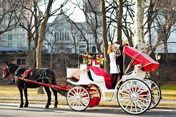 A couple shares a kiss while seated in a white and red horse-drawn carriage pulled by a black horse, in what appears to be an urban park setting with bare winter trees and a stone building visible in the background. The carriage features red upholstered seating and red trim with white spoke wheels, and is decorated with red and cream floral arrangements. The bride is wearing a white dress and the groom is in a dark suit. The shot is a wide portrait capturing the full carriage from the side.