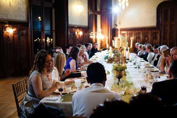 Wedding reception dinner scene inside a grand ballroom or formal dining hall featuring dark wood-paneled walls, ornate wall sconces, and warm amber lighting. Approximately 20–30 guests are seated along a long rectangular banquet-style table set with gold-toned linens, white place settings, and wine glasses. The centerpieces consist of tall candelabras with lit white tapers alongside low arrangements of green hydrangeas and white flowers. Guests are dressed in formal and semi-formal attire, including a woman in a blue dress and another in a patterned top visible in the foreground. The wide-angle shot is taken from one end of the table, giving a perspective view down its full length, with warm candlelight and wall lighting creating a dim, intimate atmosphere.