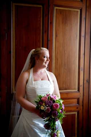 A bridal portrait taken indoors, with the bride standing in front of ornate dark wood paneling. The bride wears a white strapless A-line gown with lace detailing along the bodice and a mid-length veil with her hair pulled back. She holds a rounded bouquet featuring deep purple, mauve, and hot pink roses with trailing greenery. She is gazing to one side with a smile. The image is a medium portrait shot with warm, directional lighting that highlights the bride against the rich wood background.