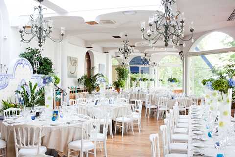 A wide shot of a fully set indoor wedding reception room with large arched windows and a glass ceiling that floods the space with natural light. The room features white Chiavari chairs, round and rectangular tables dressed in ivory or cream linens, and place settings with glassware and blue accents. Centerpieces include blue and white floral arrangements with green foliage, and multiple ornate wrought-iron chandeliers hang from the white ceiling. The decor palette is crisp white and blue with natural greenery from potted plants throughout, giving a Mediterranean or garden conservatory feel. Potential venue feature image.