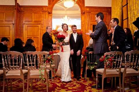 A wedding ceremony recessional taking place indoors in a formal room with warm wood-paneled walls, a crystal chandelier, and gold-toned drapery. The bride wears a fitted white lace gown with a dark sash at the waist and carries a bouquet of deep red roses, while the groom is dressed in a dark suit with a white dress shirt and boutonniere; they walk up the aisle together flanked by seated and standing guests. The ceremony chairs are chiavari-style in gold, decorated with red floral accents along the aisle, and the patterned carpet features deep red and gold tones that complement the overall decor palette. The styling is classic and formal with a bold red and gold color scheme; the shot is taken from a medium wide angle facing the couple as they exit.