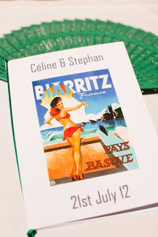 Close-up detail shot of a wedding program booklet for Céline and Stephan, dated 21st July 12, featuring a vintage-style retro travel poster illustration of Biarritz, France (Pays Basque) on the cover, depicting a woman in a red bikini. Multiple dark green card-backed programs are fanned out in the background, suggesting a Basque Country-themed wedding with a retro travel poster design aesthetic. The white booklet with grey typography is positioned in the foreground against a light surface.