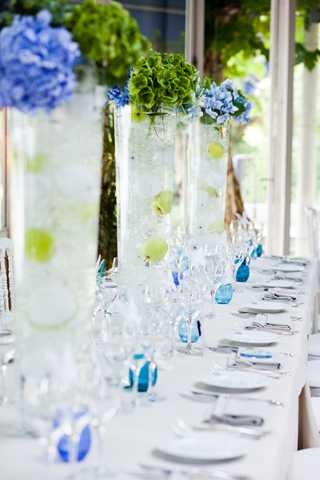 A reception table detail shot showing a long, formally set dining table with a white linen tablecloth. The centerpieces consist of tall glass cylinder vases filled with water, submerged green apples or spheres, and topped with blue hydrangeas and green hydrangeas. Small blue glass accent pieces are placed along the table between settings of white plates, polished silverware, and clear crystal stemware. The overall decor palette is blue, green, and white, creating a fresh, garden-inspired look. The setting appears to be an indoor-outdoor venue or glassed-in terrace with greenery visible through the windows in the background.