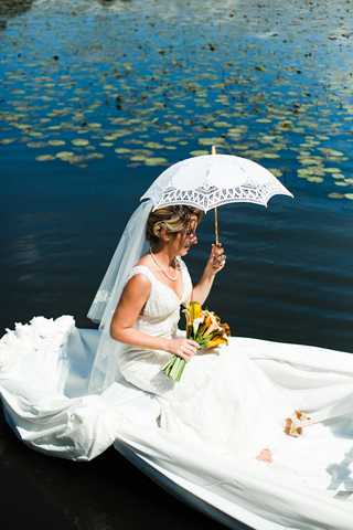 A bridal portrait taken outdoors on a white boat floating on a calm water feature with lily pads visible in the background. The bride is seated in the boat wearing a fitted white sleeveless gown with a long cathedral veil, holding a white lace-trimmed parasol in one hand and a bouquet of yellow and orange lilies in the other. Her hair is styled in a loose updo and she is wearing a pearl necklace. The shot is a medium portrait taken from slightly above, with natural bright sunlight creating strong contrast.