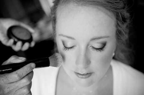 Black-and-white close-up portrait of a bride having her makeup applied during the getting-ready phase. A makeup artist's hands are visible — one holding a large powder brush to the bride's cheek and the other holding an open compact in the background. The bride has her eyes closed, hair loosely pinned up, and is wearing a white robe or top. The image has soft contrast with bright highlights on the bride's face and darker tones in the background, giving it a calm, intimate feel.