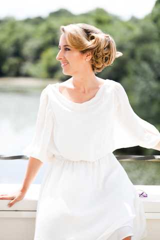 A close-up portrait of a bride standing on what appears to be a terrace or balcony railing with a body of water visible in the blurred background. She is wearing a white, loose-fitting dress with elbow-length flared sleeves and a gathered waist, giving it a relaxed, flowing silhouette. Her blonde hair is styled in an updo with soft curls pinned at the back. She is smiling and looking off to one side, with bright natural daylight illuminating the scene.