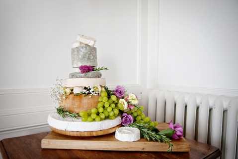 Close-up detail shot of a tiered wedding cheese cake displayed on a wooden serving board atop a rustic wooden table, set against a white-panelled interior wall with a white radiator visible to the right. The display consists of three stacked whole cheese wheels of varying sizes, decorated with fresh green grapes, purple and pink roses, white gypsophila, and sprigs of rosemary. A small round soft cheese, likely a brie or camembert, is placed in front of the board alongside additional floral and herb garnishes. The overall styling is relaxed and rustic, with a natural, garden-inspired color palette of greens, purples, and pinks.