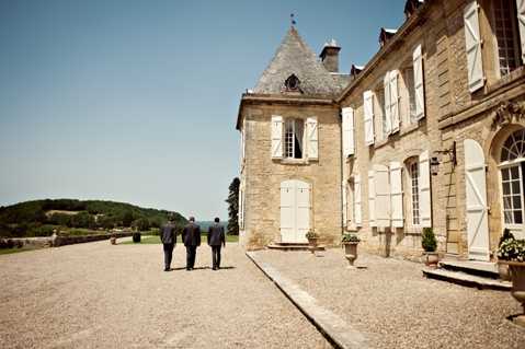 Three men dressed in dark suits walk away from the camera across a gravel courtyard alongside a French chateau with pale limestone architecture, white shuttered windows, and a slate-roofed tower. The wide shot captures the full facade of the historic manor building, with rolling hills visible in the background. The image has a slightly muted, film-like tone with soft contrast. Potential venue feature image.