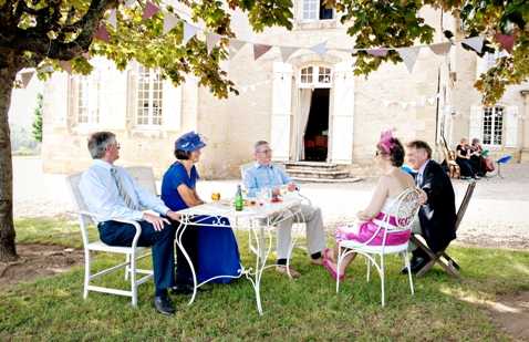 A cocktail hour or garden reception scene captured in a wide shot, showing five guests seated around a small white wrought-iron table on the lawn in front of a classic French stone manor house. Two women wear statement fascinator hats — one in royal blue dress and blue hat, another in a bright pink dress with a pink floral fascinator — while the men are dressed in suits. The table holds drinks including a green bottle and small glasses. Colorful triangle bunting is strung across the facade of the stone building in the background, and a small group of additional guests can be seen seated to the right near the entrance. The overall styling has a cheerful, British-influenced garden party feel with bold accent colors. Potential venue feature image.