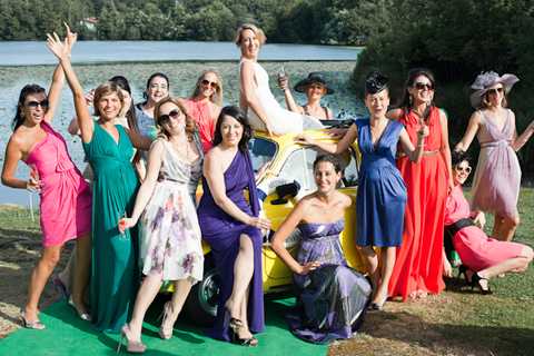 A group of approximately 14 women, including a bride in a white dress standing on top of a yellow vintage Fiat 500, pose together outdoors near a lake. The women are dressed in a wide variety of colorful formal and semi-formal attire including hot pink, coral, purple, cobalt blue, red, silver, and floral print dresses, with several wearing sunglasses and one guest in a decorative fascinator hat. The mood is celebratory and informal, with guests laughing, raising arms, and holding glasses. This appears to be a candid group portrait taken during a cocktail hour or outdoor reception, captured as a wide shot with the yellow car serving as a central prop.
