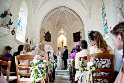 A wedding ceremony taking place inside a small historic French chapel with Gothic vaulted stone ceilings, arched windows with stained glass, and painted religious statues along the walls. The bride in a white gown and groom in a dark suit stand at the altar before an officiant, with a lit candle visible behind them. Approximately 20-30 guests are seated in traditional wooden chairs arranged in rows, with several women wearing decorative hats or fascinators. The wooden chair ends along the aisle are decorated with small bouquets of pink and white flowers tied with cream ribbon bows, adding a delicate floral detail to the classic, traditional ceremony styling. The shot is taken from the back of the chapel looking toward the altar, giving a wide perspective of the full interior.