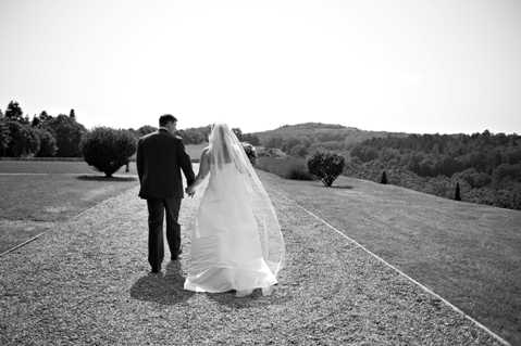 Black-and-white portrait of a bride and groom walking away from the camera hand-in-hand along a wide gravel path bordered by manicured lawns and trimmed topiary trees. The bride wears a full-length gown with a long cathedral-length veil that billows to the side in the breeze, while the groom is dressed in a dark suit. The surrounding landscape features rolling hills and dense tree lines in the background. The image is a wide mid-shot with strong contrast between the light gravel path and the darker tones of the lawn and treeline.