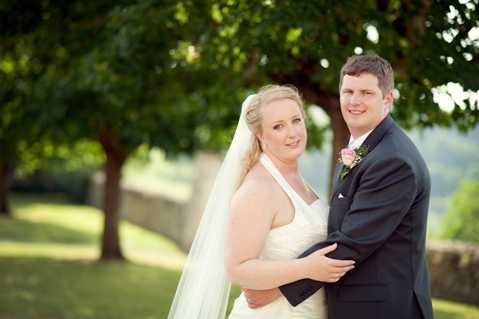 A couples portrait of a bride and groom posing outdoors on manicured grounds, likely during a post-ceremony portrait session. The bride wears a halter-neck ivory gown with a long cathedral-length veil, and her blonde hair is styled in a soft updo; the groom wears a dark charcoal suit with a light pink rose boutonniere. The pair stand close together with the groom's arm around the bride's waist, both smiling toward the camera. The shot is a mid-length portrait with a shallow depth of field, with trees and a low stone wall softly blurred in the background.