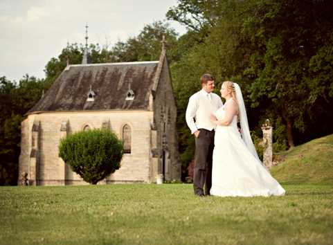 A bride and groom stand together on a lawn in front of a small stone Gothic-style chapel with a steeply pitched roof and pointed arched windows. The couple faces each other in a portrait pose, shot as a wide-angle full-length image. The bride wears a white strapless ball gown with a long cathedral-length veil, while the groom is dressed in dark trousers with a white dress shirt and light beige vest. The overall styling is classic and traditional. Potential venue feature image.