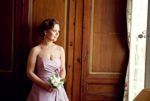 A bridesmaid stands in a portrait shot inside what appears to be a chateau or manor house, positioned beside tall wooden paneled doors with natural window light falling across her from the right. She wears a strapless dusty mauve floor-length gown with a sweetheart neckline and a gathered waist detail, and holds a small round bouquet of white roses and cream blooms with a small purple floral accent. Her dark hair is styled in an updo and she gazes off to the side toward the light source. The warm wood paneling and draped floral curtain visible at the edge of the frame suggest a classic, traditional interior setting.