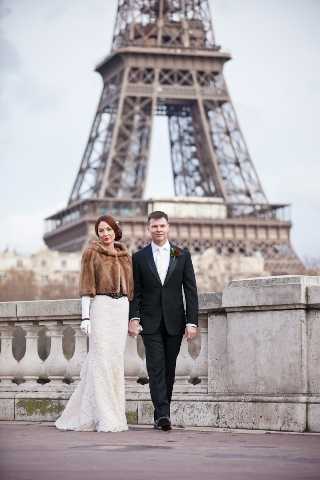 A bride and groom walk together on a stone bridge or terrace with the Eiffel Tower visible in the background, slightly out of focus. The bride wears a fitted white lace gown paired with a brown fur stole and long white gloves, while the groom is dressed in a classic black tuxedo with a white dress shirt and bow tie. The setting is outdoor Paris in what appears to be cool or winter weather, giving the portrait a classic, city editorial feel. This is a full-length couple portrait shot at a medium distance.
