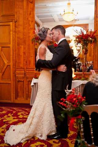 The bride and groom share their first kiss during an indoor wedding ceremony, standing at the altar embracing one another. The venue features warm wood-paneled walls, a chandelier, and a bold red and gold patterned carpet, suggesting a classic ballroom or historic hotel setting. The bride wears a fitted ivory lace gown with a small birdcage veil and fascinator, while the groom wears a black suit with a red boutonniere. Decor follows a deep red color palette, with large floral arrangements of red flowers and branches flanking the ceremony space, and a foreground arrangement of red berries and greenery visible on a nearby surface.