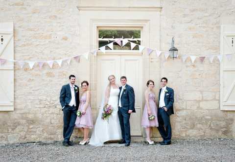 A bridal party portrait taken outdoors against the facade of a French stone building, with cream-painted double doors and wooden shutters as the backdrop. The group of six includes the bride in a white strapless ballgown with a cathedral-length veil, the groom in a navy suit with a white boutonniere, two bridesmaids in knee-length dusty lavender dresses each holding small mixed bouquets, and two groomsmen in matching navy suits. A string of pastel fabric bunting in soft pink, lavender, and cream is hung across the doorway above the group, contributing to a whimsical, vintage-inspired styling theme. The wide-format portrait shot shows all six subjects standing relaxed and smiling in a line centered on the doorway.