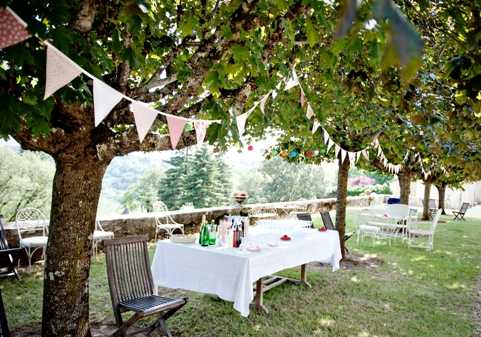 An outdoor reception or cocktail hour setup photographed in a wide shot, showing a long rectangular table draped in a white linen tablecloth set beneath mature fruit trees in a garden or orchard setting. Pink, coral, and cream fabric bunting flags are strung between the tree branches above the table. The table is set with green and clear glass bottles, small pink accents, and scattered tableware. Wooden folding chairs are placed around the table, with additional white chairs and tables visible in the background. The rustic, informal styling with fabric bunting and natural tree canopy creates a relaxed countryside aesthetic.