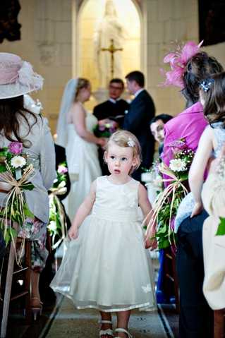 A young flower girl, approximately toddler age, stands in the center aisle of a Catholic church during a wedding ceremony, wearing a white tulle dress with a small floral hair clip, looking directly at the camera with a wide-eyed expression. In the background, the bride in a white gown and groom in a dark suit stand at the altar before a stone religious statue and crucifix. Church pew ends are decorated with mixed floral arrangements featuring pink roses, white blooms, and green foliage tied with gold and pink ribbon streamers. Guests visible on either side include women wearing decorative hats and fascinators in pink tones, with one guest in a bright fuchsia outfit. The shot is a medium portrait taken from aisle level, with the altar scene softly out of focus in the background.