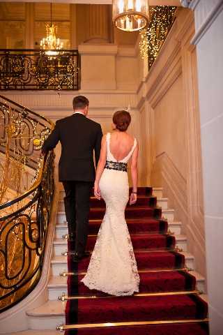 A bride and groom are photographed from behind as they walk up a grand staircase with a deep red carpet runner and ornate black and gold wrought-iron balustrade, consistent with a classic Parisian-style hotel or chateau interior. The bride wears a fitted, low-back white lace mermaid gown with a black sash detail at the waist, her hair styled in an updo, while the groom is dressed in a black tuxedo. Crystal chandeliers and warm fairy lights illuminate the upper landing, creating a warm glow. The shot is a full-length portrait taken from the base of the staircase, emphasizing the architectural details and the contrast between the bride's white gown and the red carpet. Potential venue feature image.