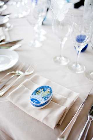 Close-up detail shot of a wedding reception place setting featuring a cream linen tablecloth and matching folded napkin placed on a white charger plate, flanked by silver cutlery. A small oval blue-and-white decorative tin, resembling a classic Camembert cheese box or similar French souvenir tin, sits on top of the napkin as a guest favor. In the background, crystal glassware including a wine glass with a cobalt blue base and a champagne flute are visible, along with additional place settings, suggesting a blue and white decor palette with a French-themed styling.