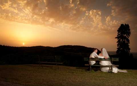 A bride and groom sit together on a wooden bench outdoors, sharing a kiss or close embrace while facing a wide landscape view at sunset. The bride wears a white gown with a long veil, and the groom is dressed in a white shirt. The couple is positioned in the lower-right area of the frame, silhouetted against a warm orange and gold sunset sky with scattered clouds. The setting appears to be an elevated outdoor terrace or lawn with a panoramic view of rolling hills. Wide shot composition with the couple as small figures against the expansive sky, emphasizing the landscape backdrop.