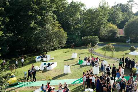 An aerial wide shot of an outdoor cocktail hour taking place on a large lawn surrounded by mature trees. Approximately 60-80 guests in formal attire are mingling across the space, which is organized into distinct lounge and standing areas. The decor includes white cocktail tables, white lounge seating with blue cushions, and white market umbrellas providing shade. A green carpet or fabric runner extends across the grass, and a vintage yellow vehicle is partially visible in the lower left corner. The overall color palette for the decor is white and blue against the green lawn setting. Potential venue feature image.