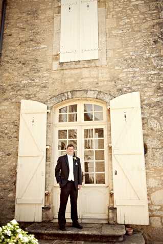 A groom stands alone in front of a traditional French stone building, positioned in a doorway framed by cream-painted wooden shutters and a glass-paned double door. He is dressed in a dark navy or black suit with a white dress shirt, white pocket square, and a small white boutonniere. The portrait is a full-length, slightly wide shot taken at ground level, with the groom centered in the composition on a low stone step. The warm, muted tones of the image give it a soft, film-like quality consistent with classic wedding photography style.