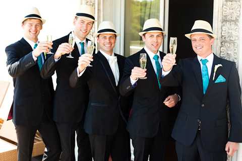 Five men — likely the groom and his groomsmen — are raising champagne flutes in a toast outdoors in front of what appears to be a modern building entrance. All five are dressed in dark navy or black suits with bright turquoise/teal neckties, and four of the five are wearing matching cream/ivory fedora-style hats. The groom or a groomsman on the far right wears a white boutonniere and a teal pocket square. The styling is cohesive with a retro-modern theme centered on the teal and black color palette. The shot is a mid-range group portrait with the men posed closely together and smiling at the camera.